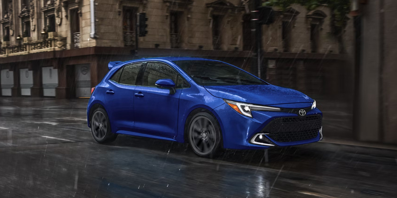 An all-new blue 2025 Toyota Corolla Hatchback driving through the rainy downtown streets. - Ed Martin Toyota Noblesville, IN