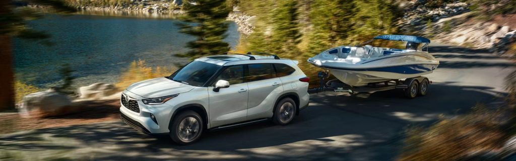 A white SUV towing a boat on a scenic road near a lake, surrounded by trees.