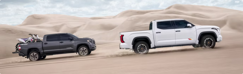 Gray Toyota Tundra carrying dirt bikes beside a white Toyota Tundra on desert sand dunes
