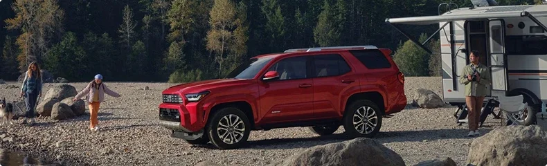Red Toyota 4Runner parked on a rocky lakeshore beside a travel trailer as a family walks by