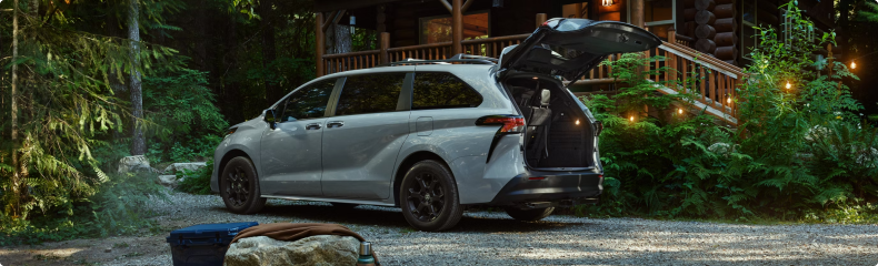 White Toyota Sienna with its liftgate open on a gravel drive outside a log cabin