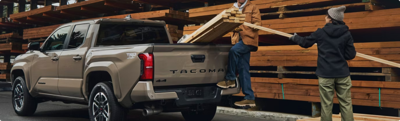 Two people loading lumber into the bed of a tan Toyota Tacoma at a timber yard